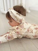 Zbliżenie na dziewczynkę leżącą na brzuszku na drewnianej podłodze, ubraną w kwieciste body z widocznymi falbankami i dopasowaną opaską.