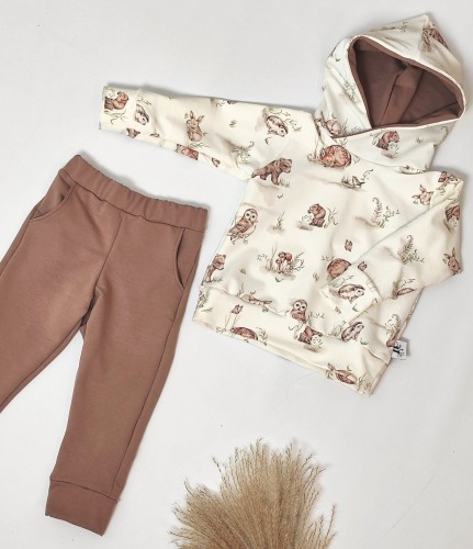 Komplet ubranek dziecięcych handmade: bluza z kapturem z motywem leśnym oraz gładkie, brązowe spodnie dresowe z kieszeniami, leżące płasko na jasnym tle.