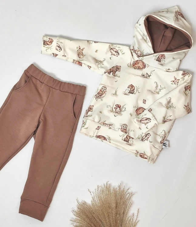 Komplet ubranek dziecięcych handmade: bluza z kapturem z motywem leśnym oraz gładkie, brązowe spodnie dresowe z kieszeniami, leżące płasko na jasnym tle.