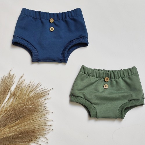 Dwie pary dziecięcych retroshortów handmade w kolorze ciemnoniebieskim i oliwkowym z ozdobnymi drewnianymi guzikami na tle suszonych traw
