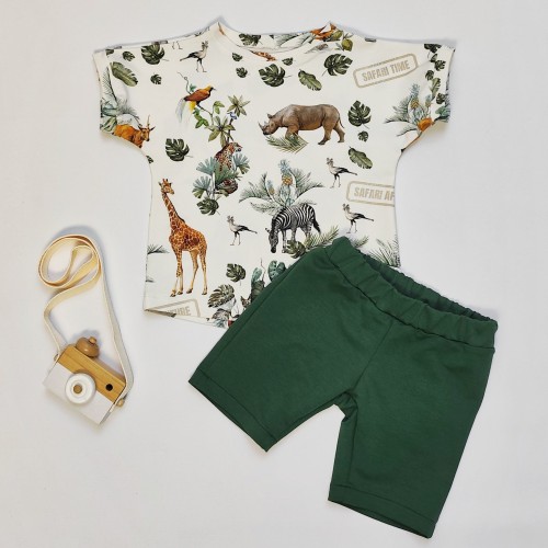 Zdjęcie przedstawia leżący komplet ubranek handmade dla dzieci na białym tle. T-shirt z motywem safari (żyrafa, słoń, lew, zebra) i zielone krótkie spodenki. Obok leży drewniany aparat fotograficzny.