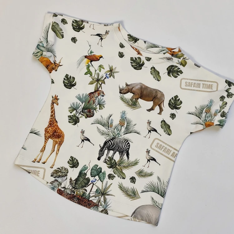 Zbliżenie na t-shirt handmade dla dzieci z motywem safari. Na zdjęciu widać fragmenty wzoru z żyrafą, słoniem, lwem, zebrą i liśćmi monstery.